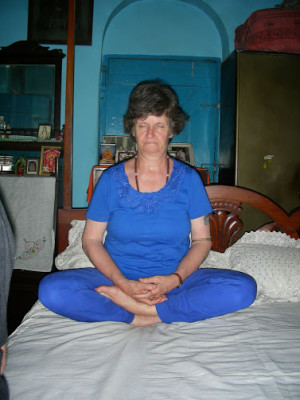 Brindey seated in half-lotus pose on a bed.