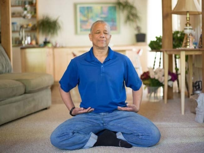 Man meditating in a living room on a cushion, relaxed expression on his face.